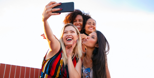 Cuatro mujeres jóvenes sonriendo y posando mientras se toman una selfie al aire libre, con cielo despejado de fondo